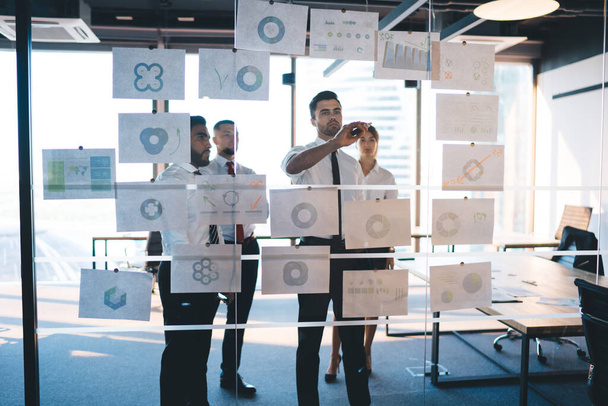 Diversity analysts in formal clothing creating marketing plan for corporate productivity, successful male and female experts brainstorming during working process with revenue infographics in office - Photo, Image