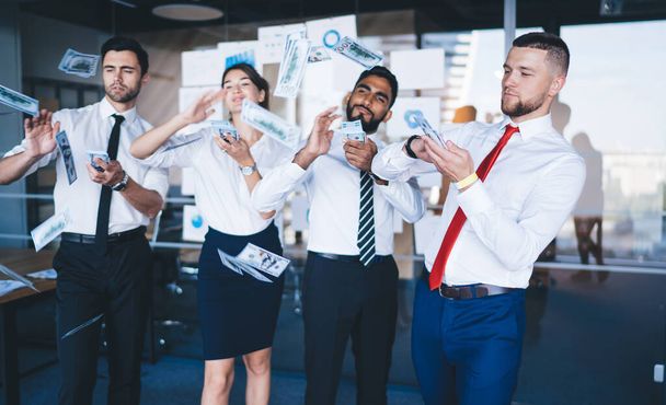 Group of well dressed elegant colleagues throwing money bills in modern well lit office space with glass wall behind - Photo, image