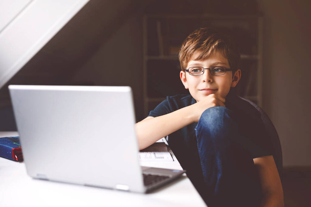 Chłopiec w okularach uczący się w domu na laptopie do szkoły. Urocze dziecko odrabiające lekcje i korzystające z notebooka i nowoczesnych gadżetów. Koncepcja szkolnictwa domowego. - Zdjęcie, obraz