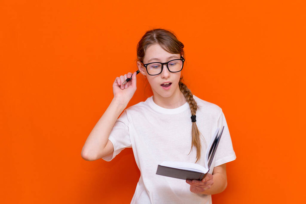 Aufgeregtes Nachdenken, wenn man auf ein junges Mädchen mit Brille, weißem T-Shirt und Stift blickt und auf ein Notizbuch auf orangefarbenem Studiohintergrund blickt.. - Foto, Bild