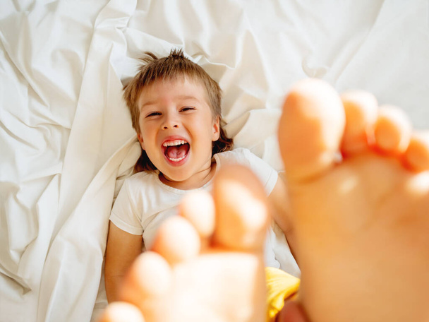 Little boy is lying upside down on the bed and laughing happily. Joyful toddler. Playful child shows his feet in the air. Sunny morning in cozy home. - Zdjęcie, obraz
