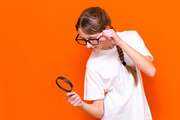 Attractive young girl in glasses looks through a magnifier on a bright orange background. - Фото, зображення