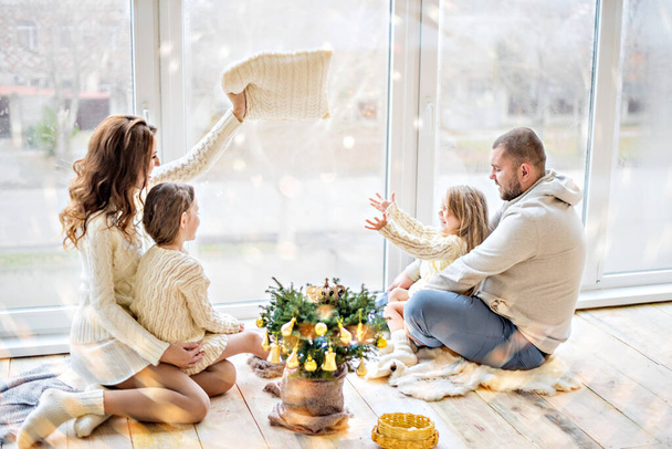 A beautiful young couple family and their two daughters are having fun celebrating New Year. Christmas tree in a pot, large panoramic window and festive lights. Copy space, card bokeh. Winter resting - Foto, Imagem