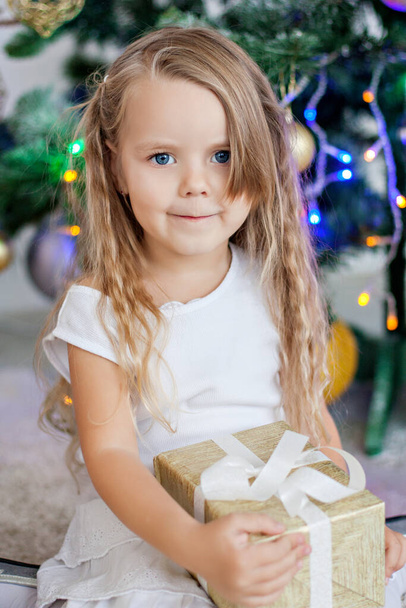 Close-up portrait of a little blonde girl in a white dress, holding a gift near the Christmas tree with a garland of lights. New Year holiday by the fireplace at home. Happy child have fun. Copy space - Foto, Imagen