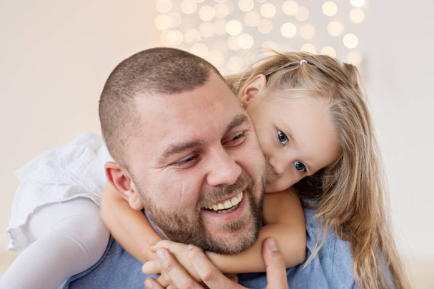 A close-up portrait of a happy father and a little girl against a yellow bokeh white background. A young man with a beard plays and hugs his daughter. New Year mood, Christmas garland. Copy space card - Φωτογραφία, εικόνα