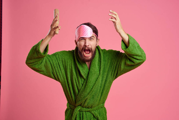 Retrato de un hombre con una máscara de sueño rosa y un rodillo de madera emociones modelo de irritabilidad bata verde - Foto, imagen