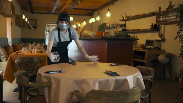 Kelnerka, nosząca maskę chirurgiczną i rękawiczki, nakrywająca do stołu w restauracji. Kobieta właściciel ponownie otworzyć małą firmę po zamknięciu, podczas epidemii koronawirusu. - Materiał filmowy, wideo