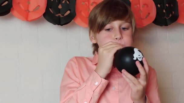 Un chico rubio sonriendo Inflando el globo negro con un fantasma, prepárate para Halloween en casa, celebra Halloween en casa con decoraciones festivas - Metraje, vídeo