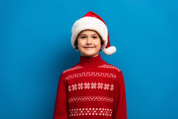 happy boy in santa hat and red sweater looking at camera on blue - Φωτογραφία, εικόνα