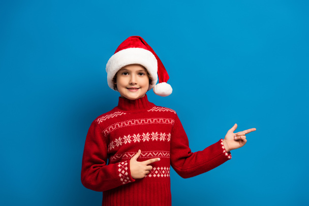 joyful boy in santa hat and red sweater pointing with fingers on blue - Photo, Image