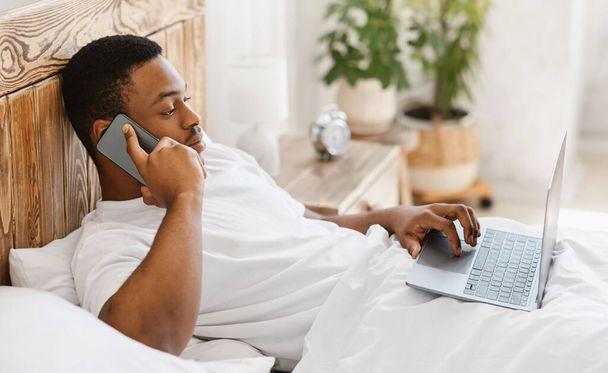 Africain homme parlant sur téléphone portable travaillant sur ordinateur portable au lit - Photo, image