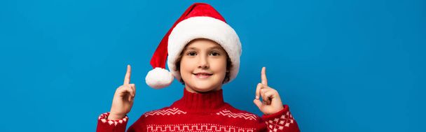horizontal image of joyful kid in santa hat and red sweater pointing with fingers while looking at camera on blue - Fotó, kép