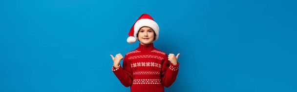 Panoramaaufnahme eines fröhlichen Kindes mit Weihnachtsmannmütze und rotem Pullover, das die Daumen nach oben zeigt, während es isoliert auf blau in die Kamera blickt - Foto, Bild
