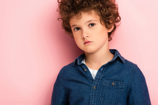 serious and curly boy in denim shirt looking at camera on pink  - Foto, Bild