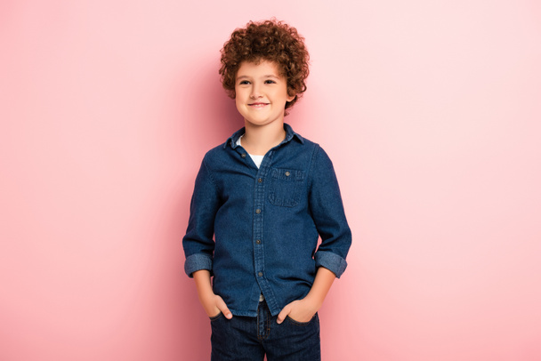 tevreden en krullende jongen in denim shirt staan met de handen in zakken op roze  - Foto, afbeelding