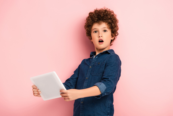chocado y rizado niño sosteniendo tableta digital en rosa  - Foto, imagen