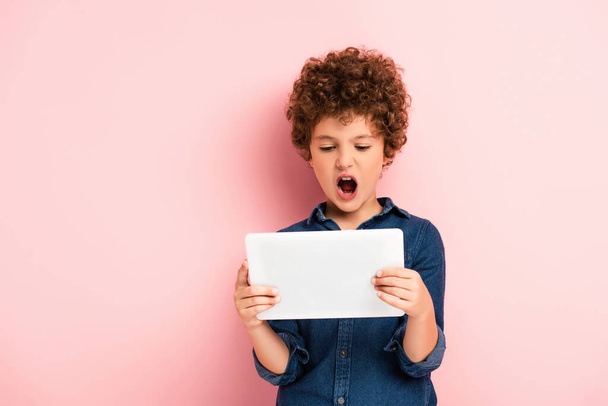 θυμωμένο και σγουρό παιδί ουρλιάζοντας και κοιτάζοντας ψηφιακή ταμπλέτα σε ροζ  - Φωτογραφία, εικόνα