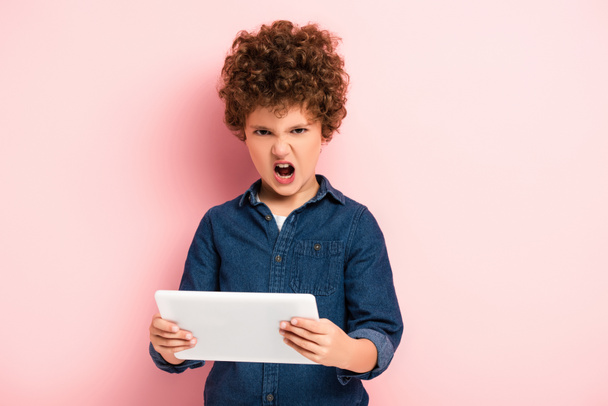 angry and curly kid screaming and holding digital tablet on pink  - Foto, Imagen