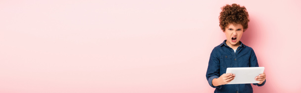 plano panorámico de niño rizado enojado gritando y sosteniendo la tableta digital en rosa  - Foto, Imagen