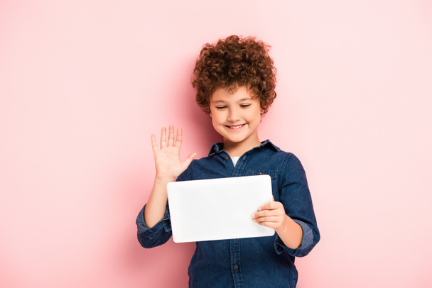 σγουρό παιδί κουνώντας το χέρι, ενώ χρησιμοποιώντας ψηφιακή ταμπλέτα κατά τη διάρκεια της κλήσης βίντεο σε ροζ  - Φωτογραφία, εικόνα