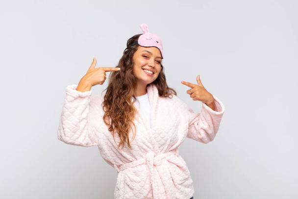 young pretty woman smiling confidently pointing to own broad smile, positive, relaxed, satisfied attitude wearing pajama - Foto, Bild
