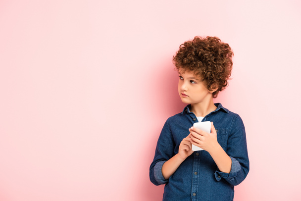 curly kid in denim shirt holding smartphone on pink - Photo, Image