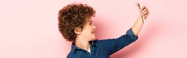 panoramic crop of curly kid in denim shirt taking selfie on smartphone on pink - Fotoğraf, Görsel