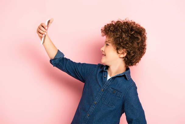 side view of curly kid in denim shirt taking selfie on smartphone on pink - Foto, Imagen