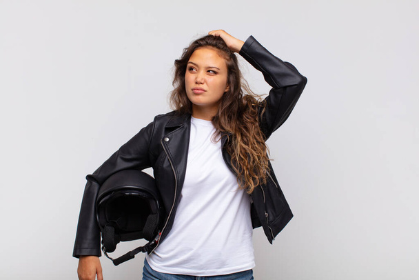young woman motorbike rider feeling puzzled and confused, scratching head and looking to the side - Foto, Imagen