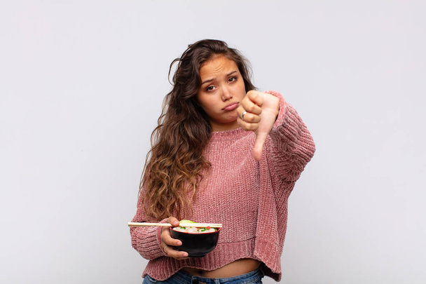 young pretty woman with noodles feeling cross, angry, annoyed, disappointed or displeased, showing thumbs down with a serious look - Foto, afbeelding