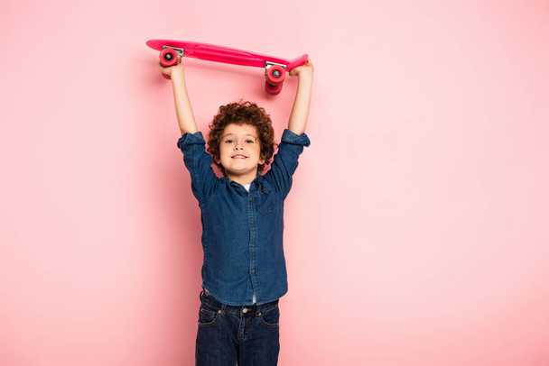 curly boy holding penny board above head on pink  - Fotó, kép