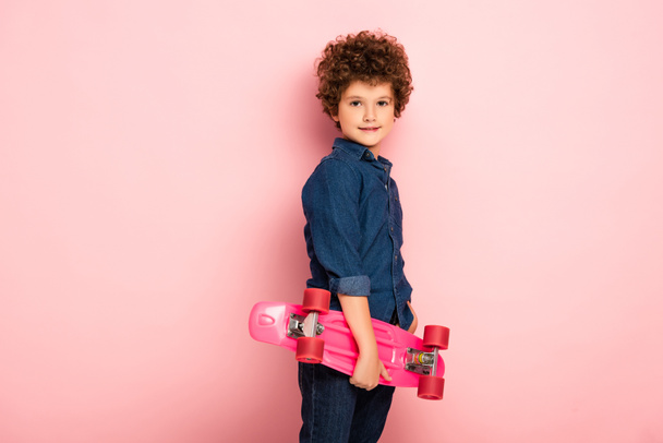 krullend jongen holding penny board en glimlachen op roze  - Foto, afbeelding