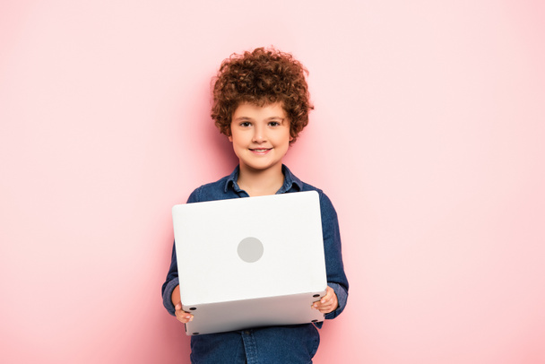 alegre y rizado chico celebración de la computadora portátil en rosa  - Foto, Imagen