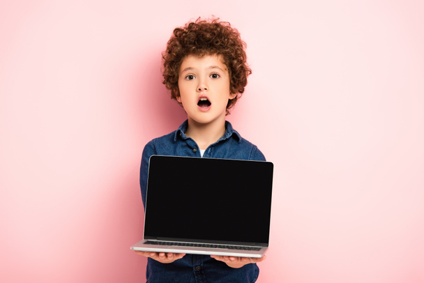 šokovaný a kudrnatý chlapec drží notebook s prázdnou obrazovkou na růžové  - Fotografie, Obrázek