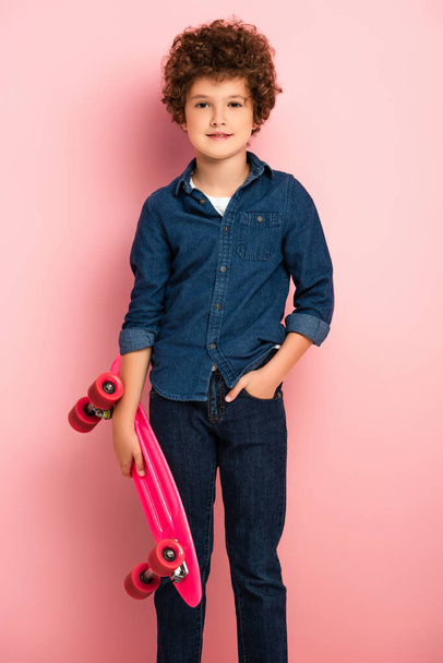 curly boy holding penny board and standing with hand in pocket on pink  - Фото, изображение