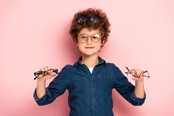 joyful boy holding bunch of eyeglasses while looking at camera on pink   - 写真・画像