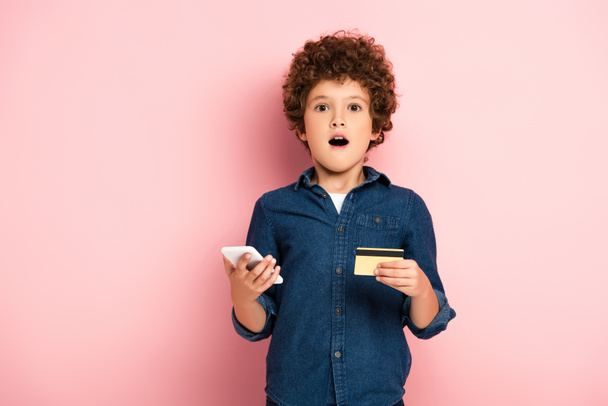 menino chocado segurando smartphone e cartão de crédito enquanto compras on-line em rosa  - Foto, Imagem
