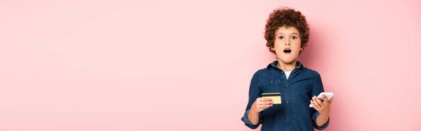 panoramatický koncept šokovaného chlapce držícího smartphone a kreditní kartu při on-line nakupování na růžové  - Fotografie, Obrázek
