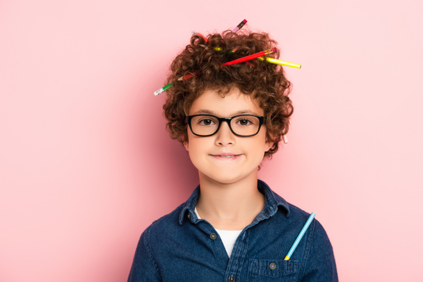 zufriedenes Kind in Brille mit Buntstiften im lockigen Haar auf rosa  - Foto, Bild
