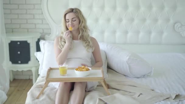 Smiling pregnant woman eating chips and caressing belly. Camera approaches to happy Caucasian woman chewing unhealthy food during pregnancy. Potential harm to fetus. - Záběry, video