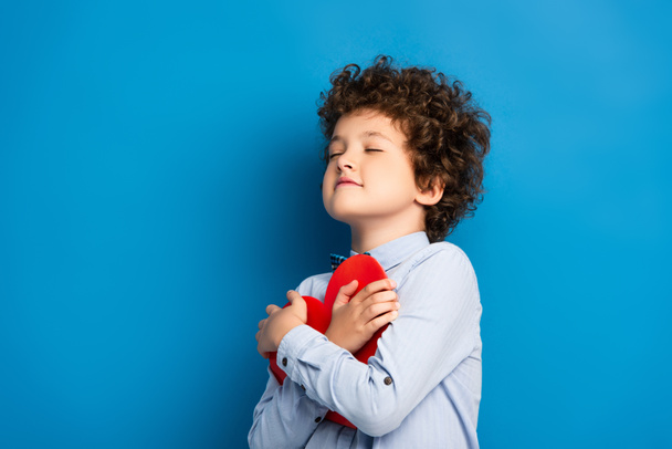 joyful kid with closed eyes holding red paper cut heart on blue - Foto, afbeelding