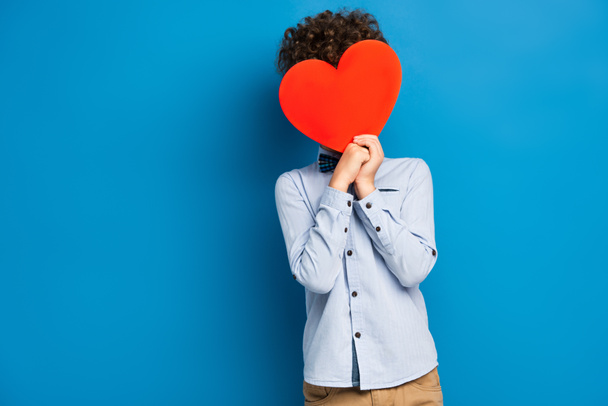 curly boy covering face while holding red paper cut heart on blue - Φωτογραφία, εικόνα