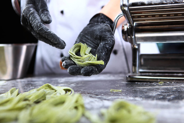The chef rolls freshly prepared pasta into nests. Dough with spinach. Healthy food. Unrecognizable person. Photo on a dark background. Hands in protective gloves.Bottom view.Close up. - Photo, Image