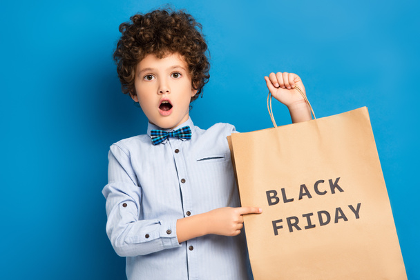 geschokt en krullend jongetje wijzend met vinger naar boodschappentas met zwarte vrijdag belettering op blauw  - Foto, afbeelding