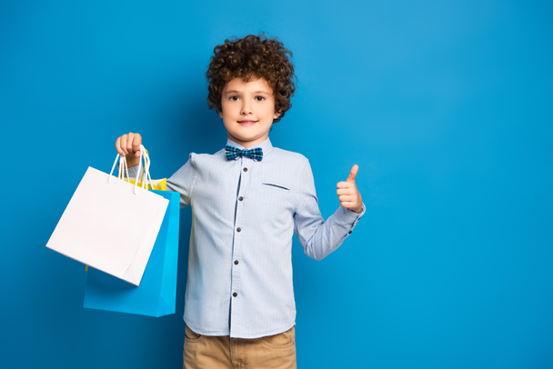 vrolijk en krullend jongen houden boodschappentassen en tonen duim omhoog op blauw  - Foto, afbeelding