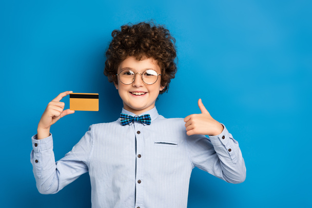 χαρούμενο αγόρι με γυαλιά που κρατάει πιστωτική κάρτα και δείχνει τον αντίχειρα πάνω στο μπλε - Φωτογραφία, εικόνα