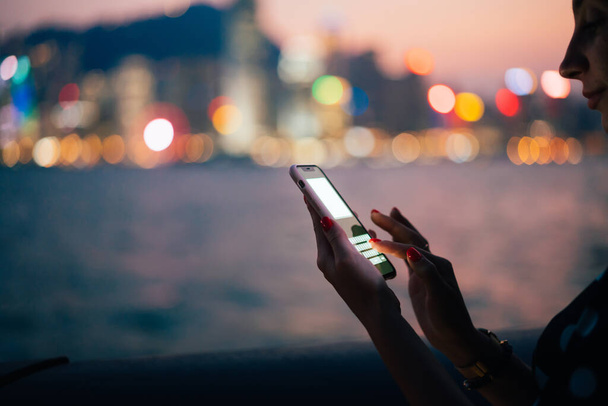 Side view of crop unrecognizable female using modern smartphone in plastic case with messenger mock up on blurred background of night river and bokeh city lights - Foto, afbeelding