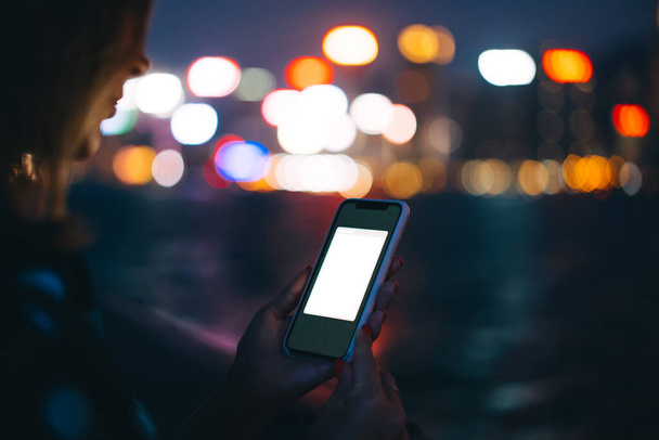 Crop unrecognizable female using cellphone in plastic case with application mock up on blurred background of night river and bokeh city lights - Foto, afbeelding