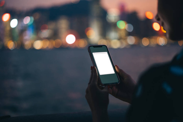 Ritaglia femmina irriconoscibile utilizzando il moderno dispositivo telefonico in custodia di plastica con applicazione modellare su sfondo sfocato di notte fiume e bokeh luci della città - Foto, immagini
