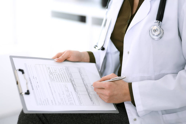 Woman doctor writing something at clipboard while sitting at the chair, close-up. Therapist at work filling up medication history records. Medicine and healthcare concept - Photo, image